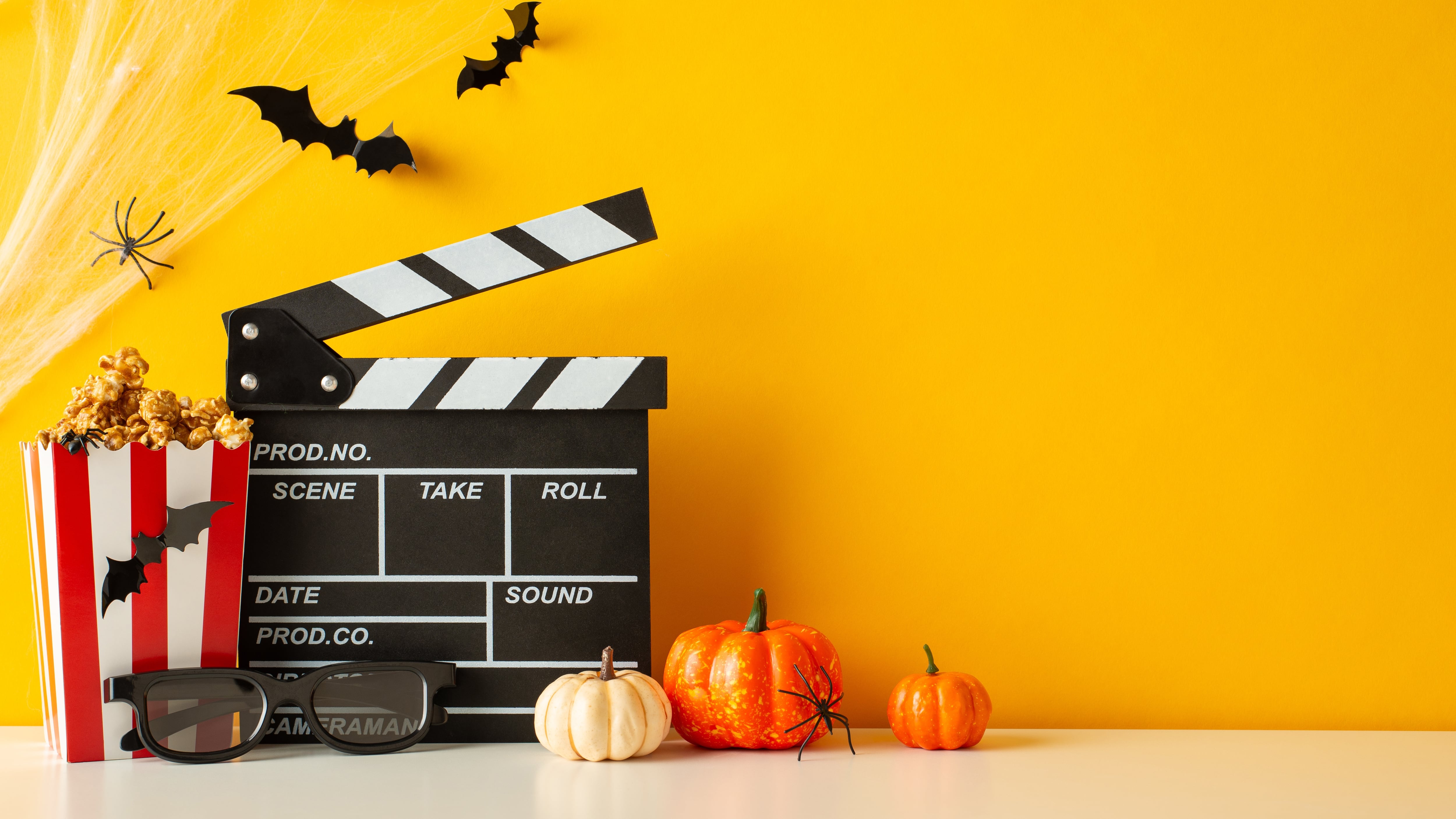 Side view shot features a table adorned with pumpkins, insects, 3D glasses, clapperboard and popcorn boxes on yellow wall background, spooky movie promo
